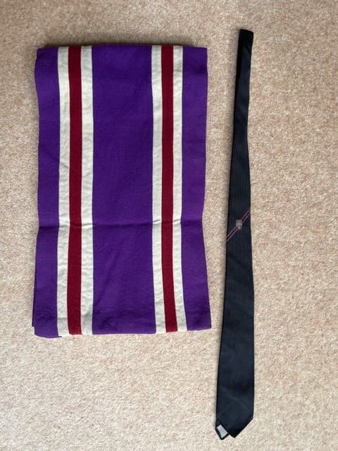 A Loughborough University branded scarf and tie. The scarf is striped purple, white and pink and the tie is black with the Loughborough University logo with purple and white.